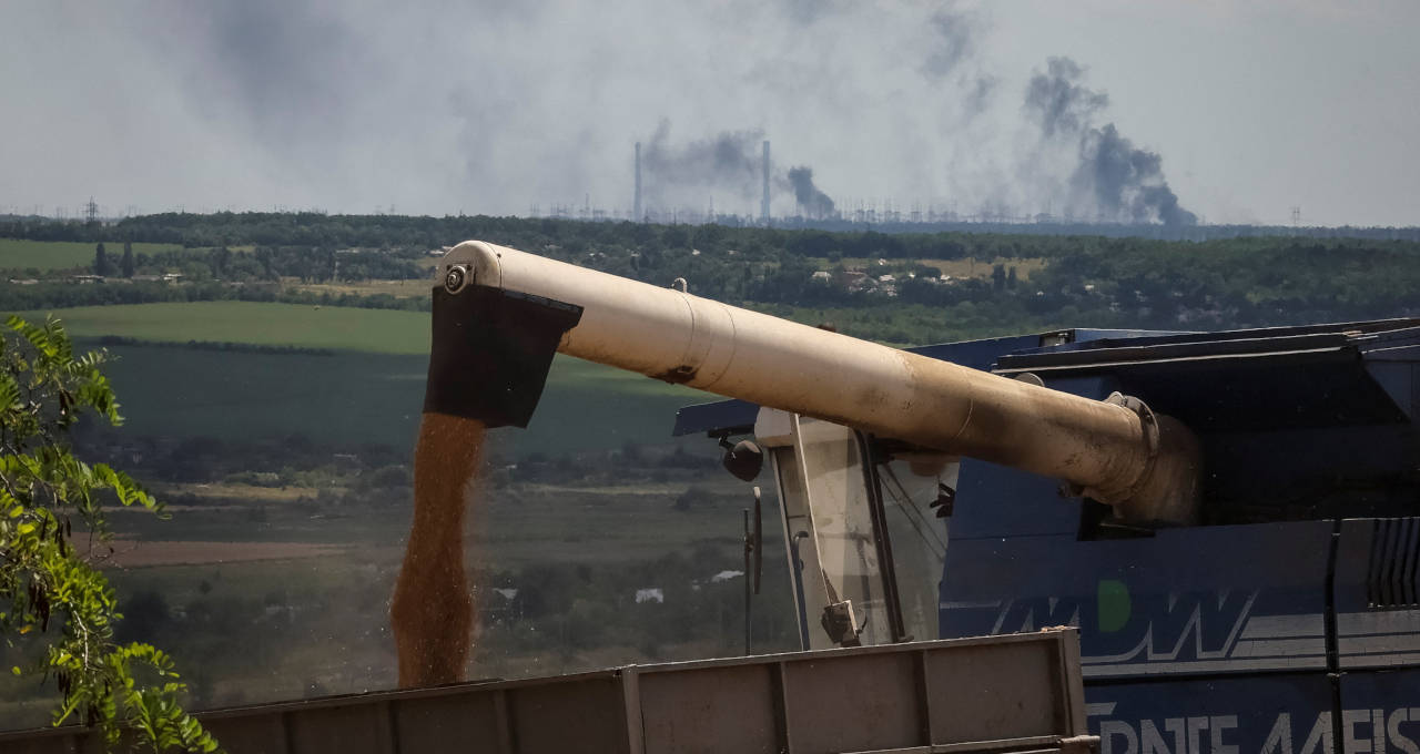Agricultores colhem trigo na região ucraniana de Donbas enquanto usina de energia queima ao fundo, após ataque de artilharia durante invasão russa na Ucrânia